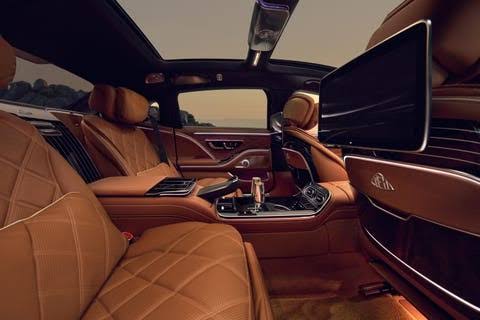 The rear cabin of a Mercedes saloon at sunset, quilted tan leather and an open panoramic roof.